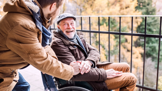 A young man patting an older man in a wheelchair on the arm