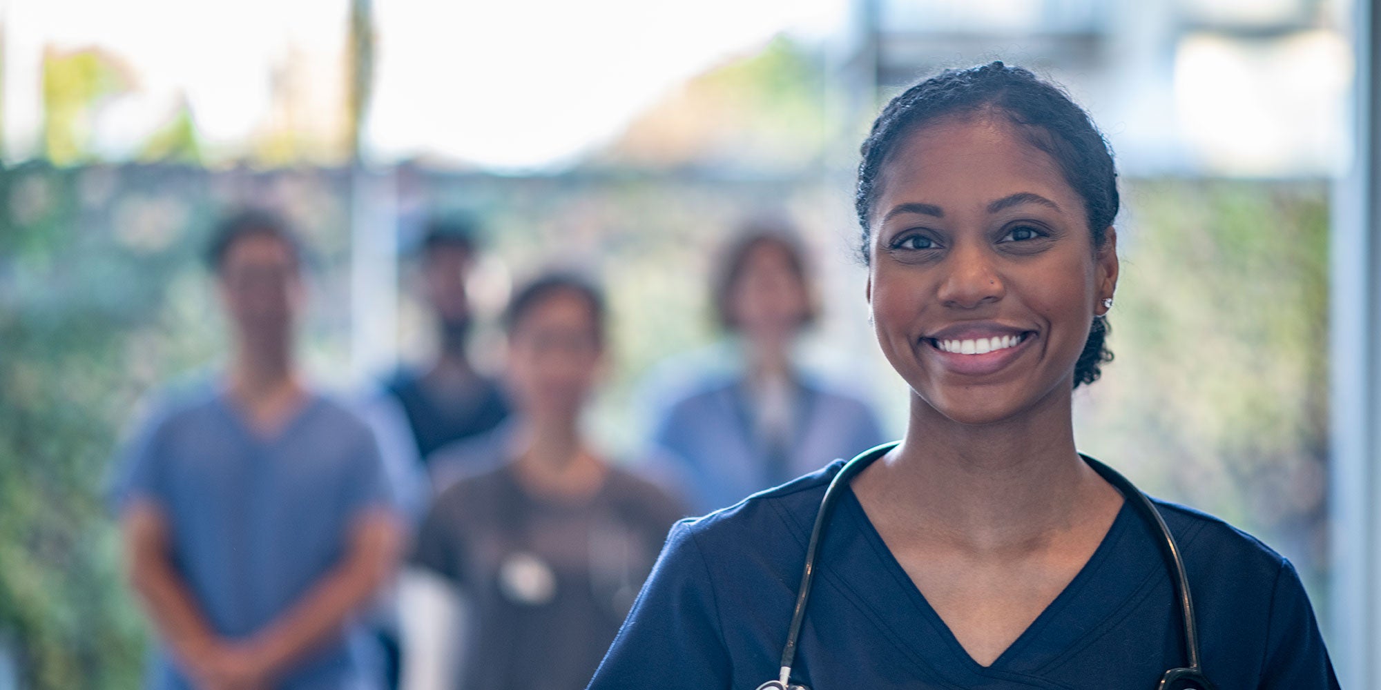 A medical resident stands at the forefront of the image smiling. She's wearing blue scrubs and a group of other medical professionals stand behind her.