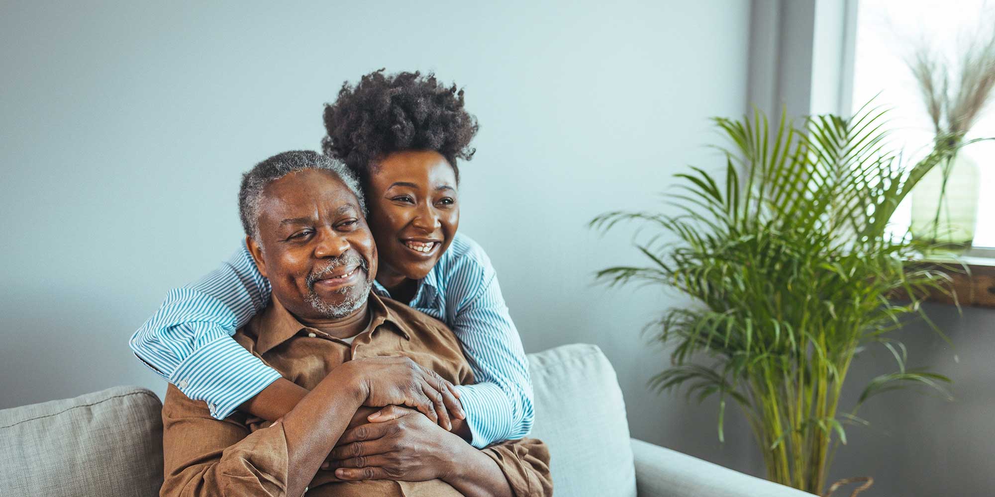 A daughter hugs her dad from behind. He's sitting on the couch. Next to them a plant is illuminated by sunlight coming through a window.