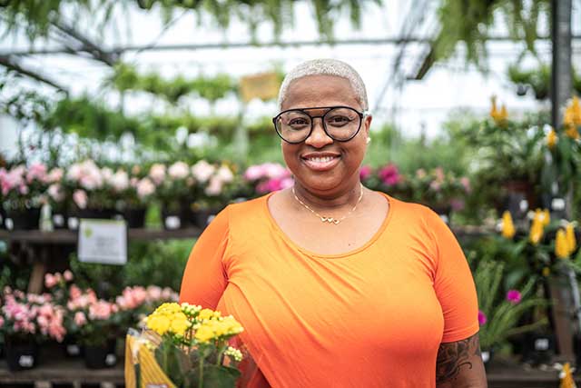 A woman in an orange shirt stands in a greenhouse smiling. She's holding a bundle of yellow flowers.