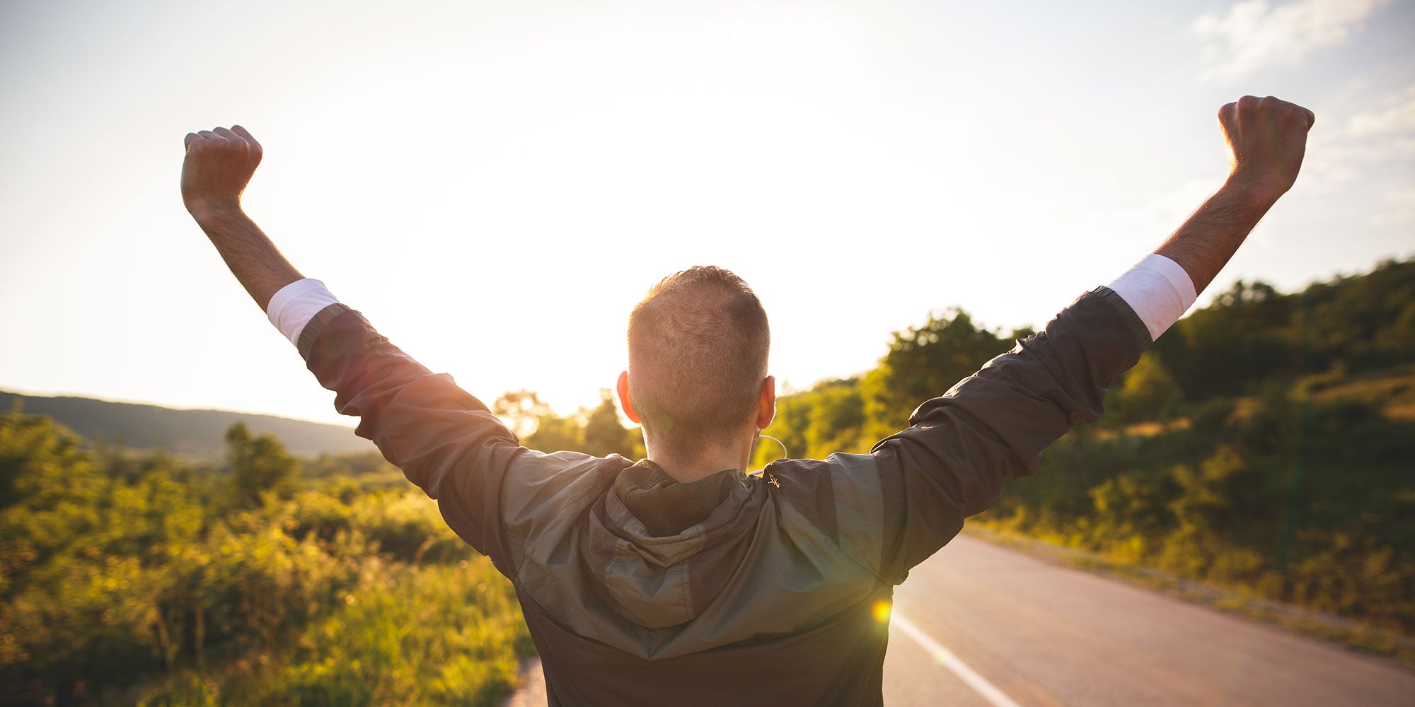 A man raises his arms in joyous celebration. He's standing on an empty road as the sun sets in the foreground.