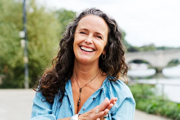 A woman joyfully touches her hands together along a river walk. She's wearing a blue, flowy shirt. There are green trees and shrubbery in the background.