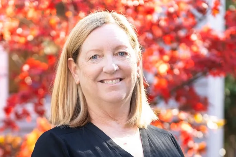 A woman named Melinda stands in front of a bright red tree in autumn. She's wearing a black blouse and is smiling gratefully.