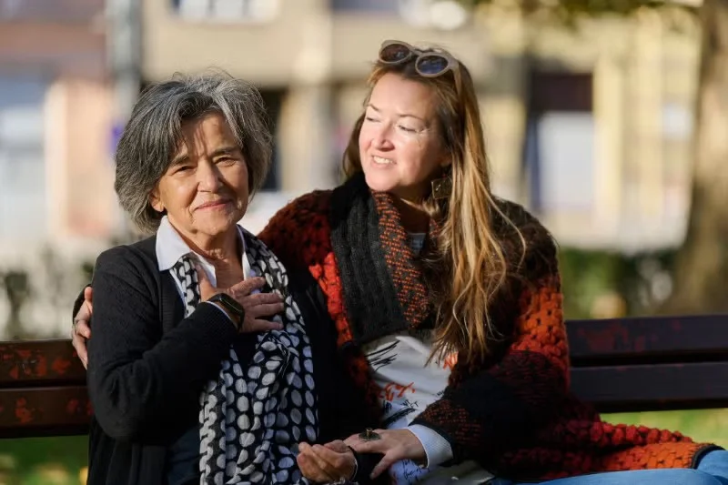 A young adult woman wraps her arm around her mother on a park bench. They're both wearing sweaters, and the weather appears brisk.