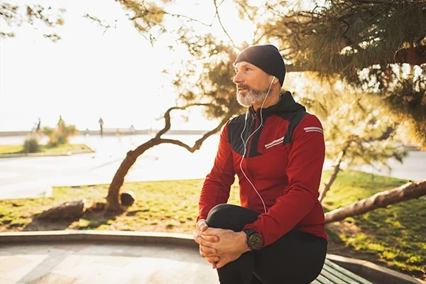 A man in exercise clothes clutches his knee while catching his breath. He is on a run in crisp weather.