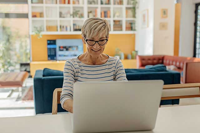 An aged woman with white hair types on her computer. She's wearing a white and black striped shirt.
