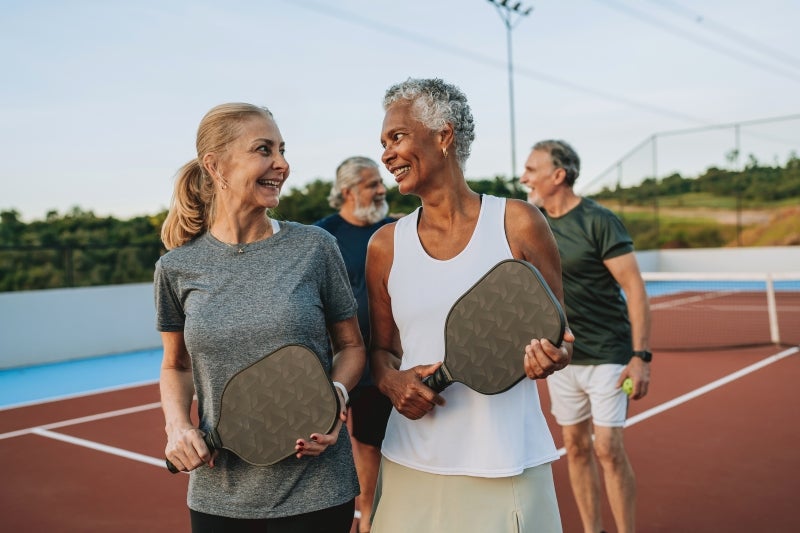 two ladies hanging on to pickleball paddles smiling at each other