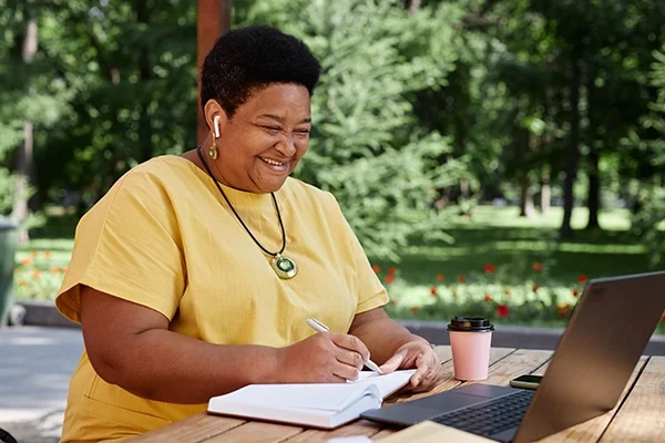 A woman sits at her laptop computer and takes notes in a journal