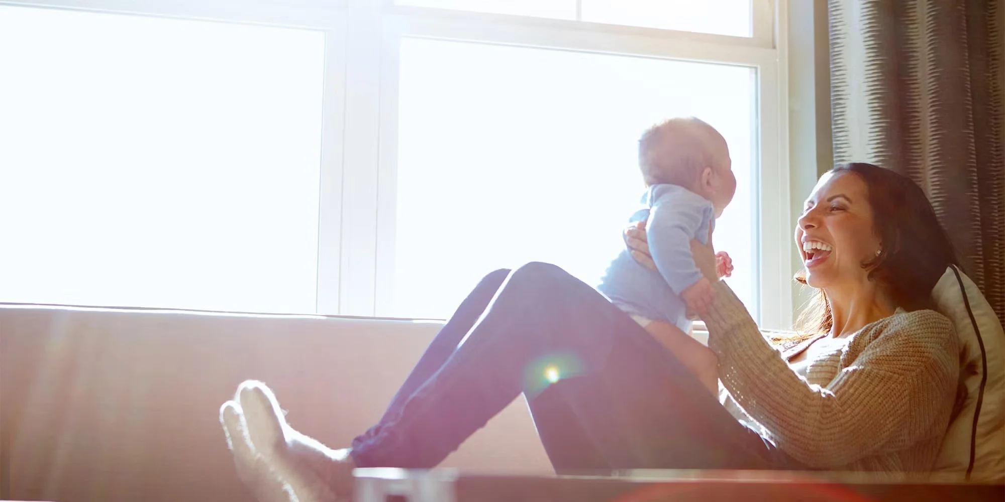 A young mother holds her baby up on a couch. Sunlight beams in through the window.