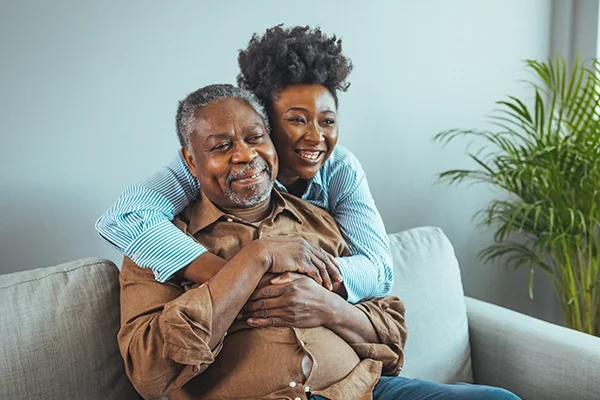 A woman wraps her arms around her father from behind where he's sitting