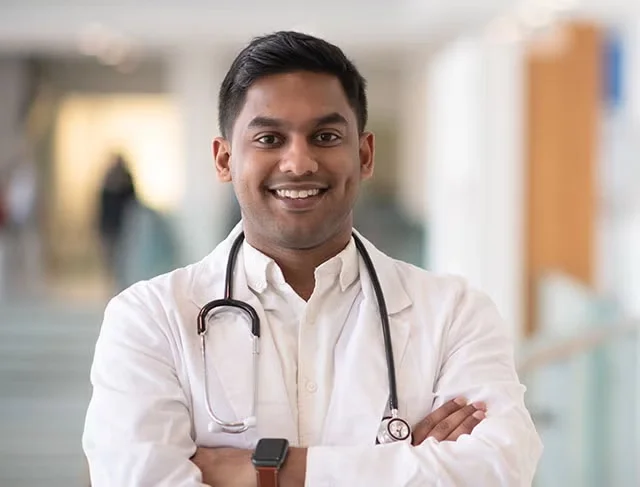 A doctor stands smiling with his arms crossed in a hospital hallway. His stethoscope is draped around his neck.