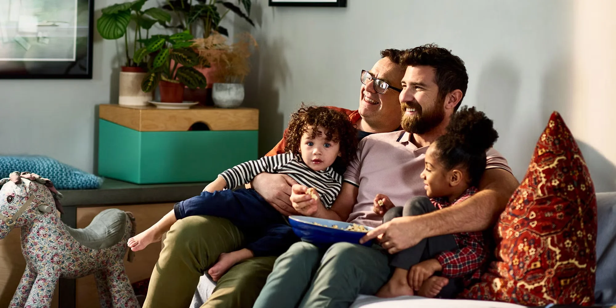 Two men cuddle their children together on a couch