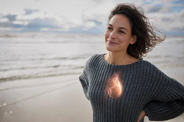 A woman stands on a windy beach with her hands on her hips. There is a graphic of a heart laying over where her heart would be on her chest.