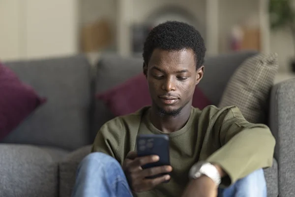 A man sits on the ground leaning on a couch and looks at his phone