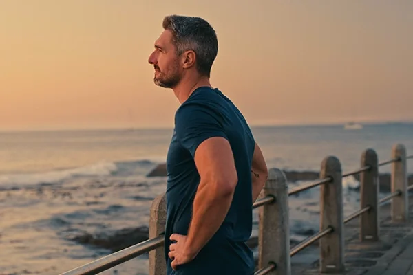 A fit middle-aged man stands on a pier with his hands on his hips. He's peering out thoughtfully over the lake.