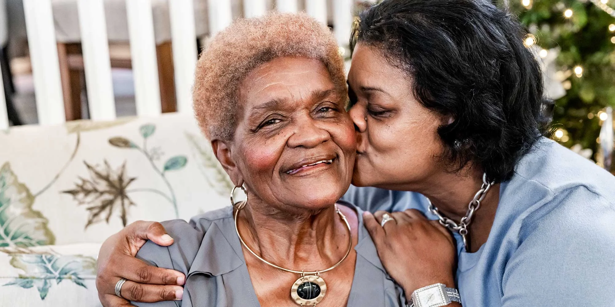 A senior woman is being kissed on the cheek by her daughter. They're sitting at the bottom of a front porch area.