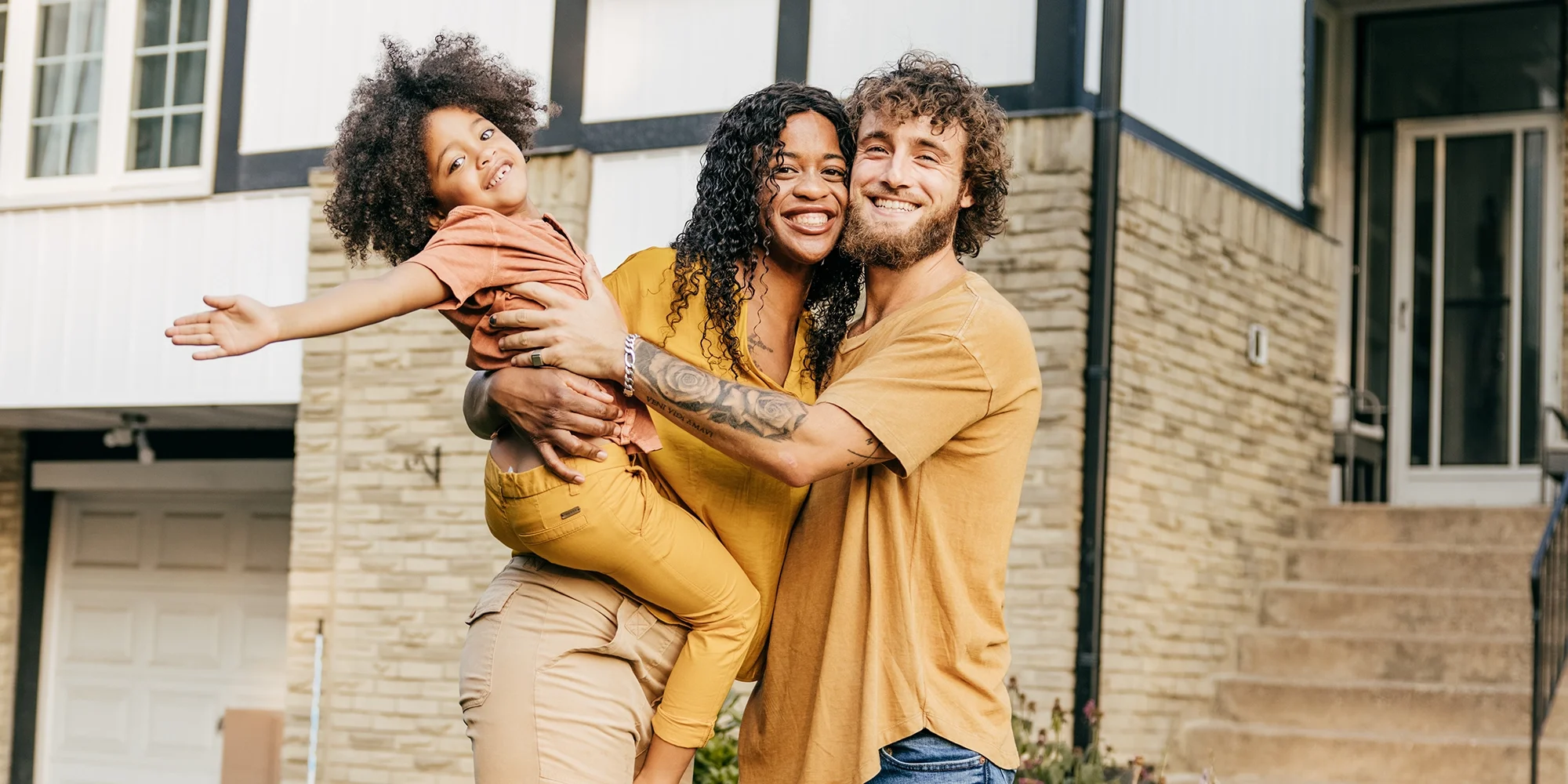 A cute family standing outside their home. The young boy is up in his mother's arms, happily gesturing outwardly.