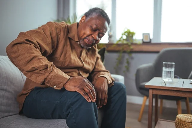 An aging man sitting on a couch winces in pain as he clutches his knee.