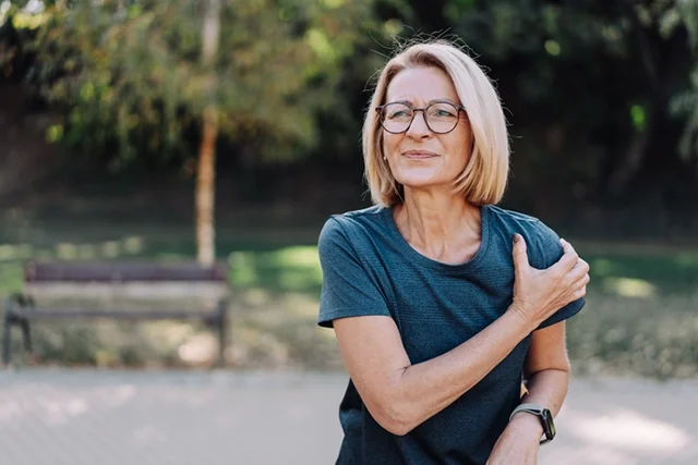 A middle-aged woman walking in a park grabs her shoulder in pain.