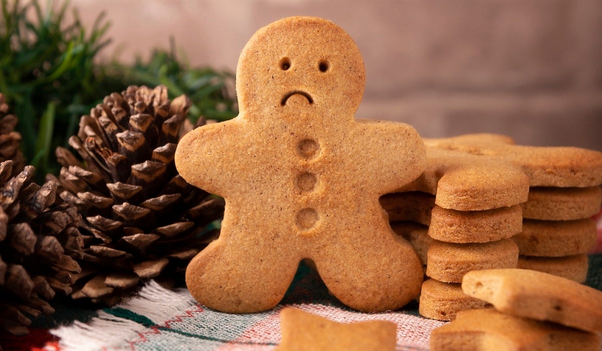 Gingerbread cookie with a sad face, next to a stack of other cookies set on a table with a pinecone and greenery for decor.
