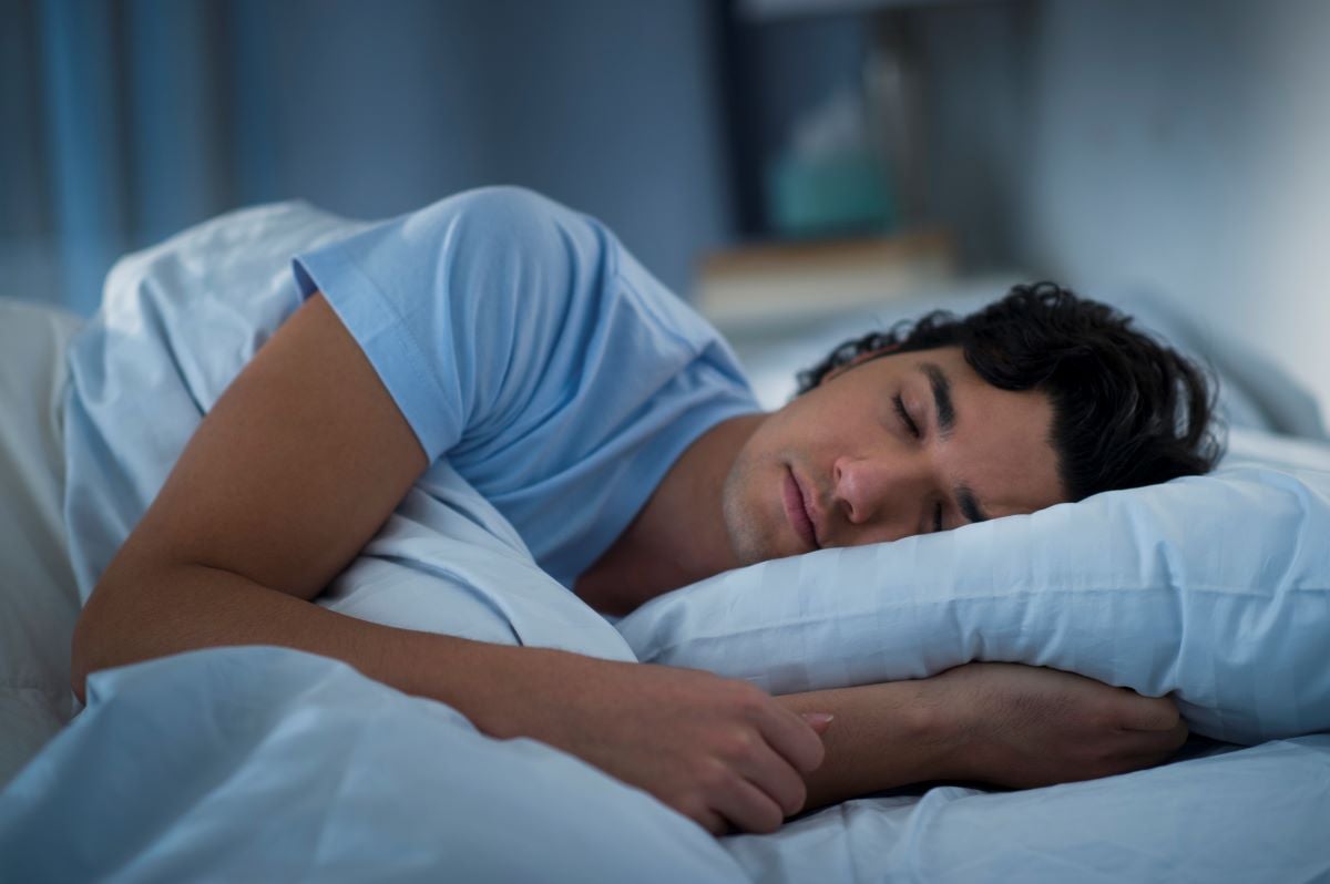 Caucasian man sleeping in bed with white sheets surrounded by dark, calm room.