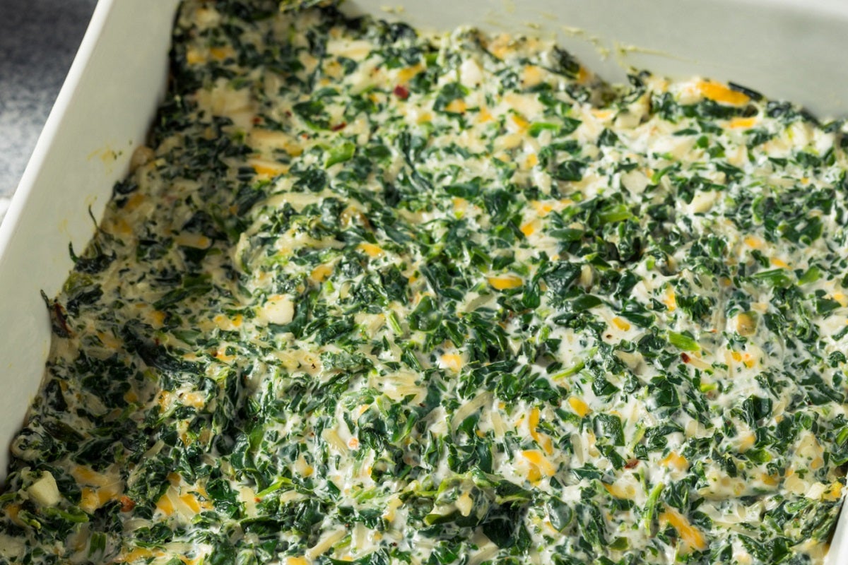 Spinach, white bean, and artichoke dip mixture in a white ceramic dish and placed on a grey table covered in a striped cloth.